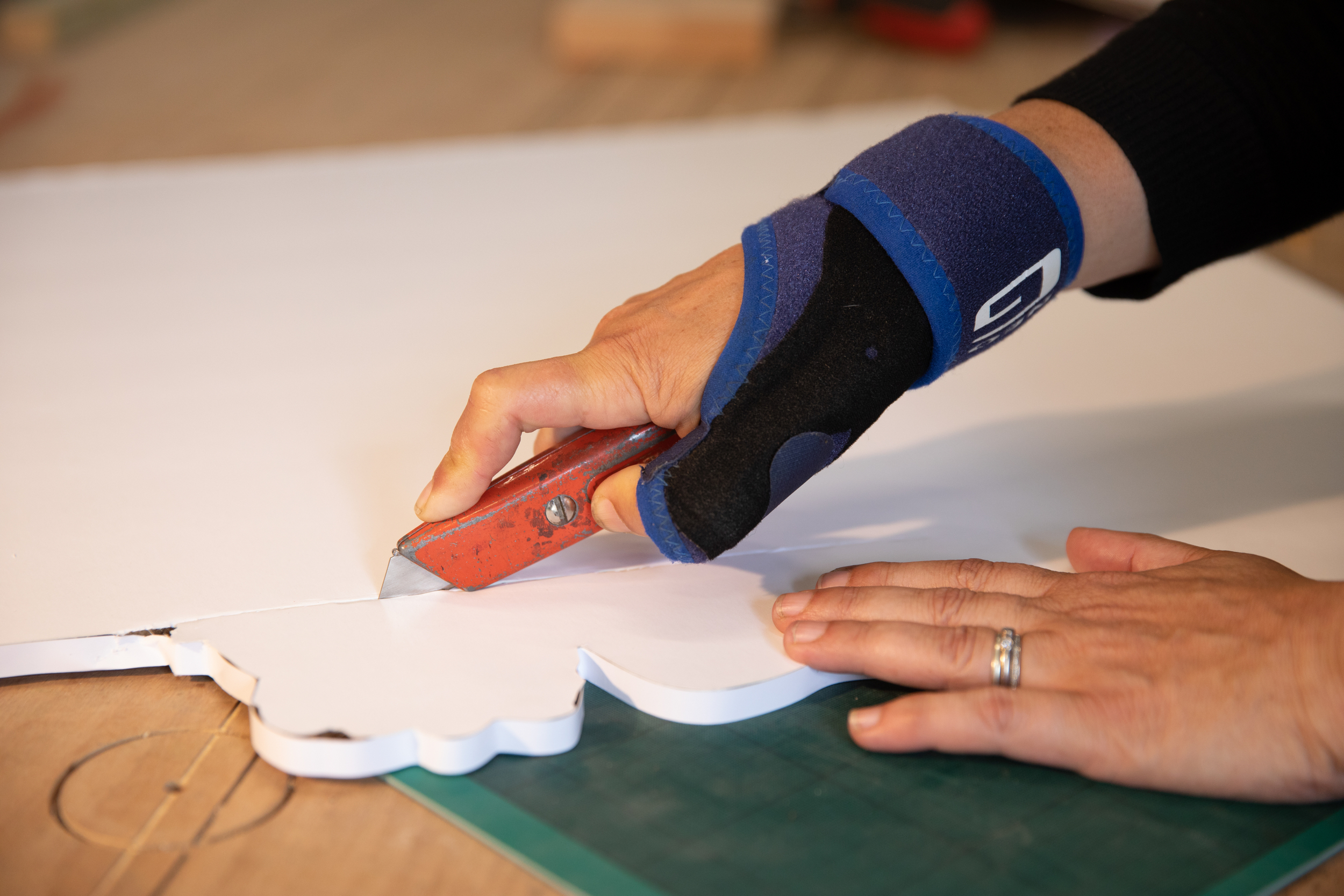 A hand with a wrist splint cuts a sheet of thick white card with a craft knife
