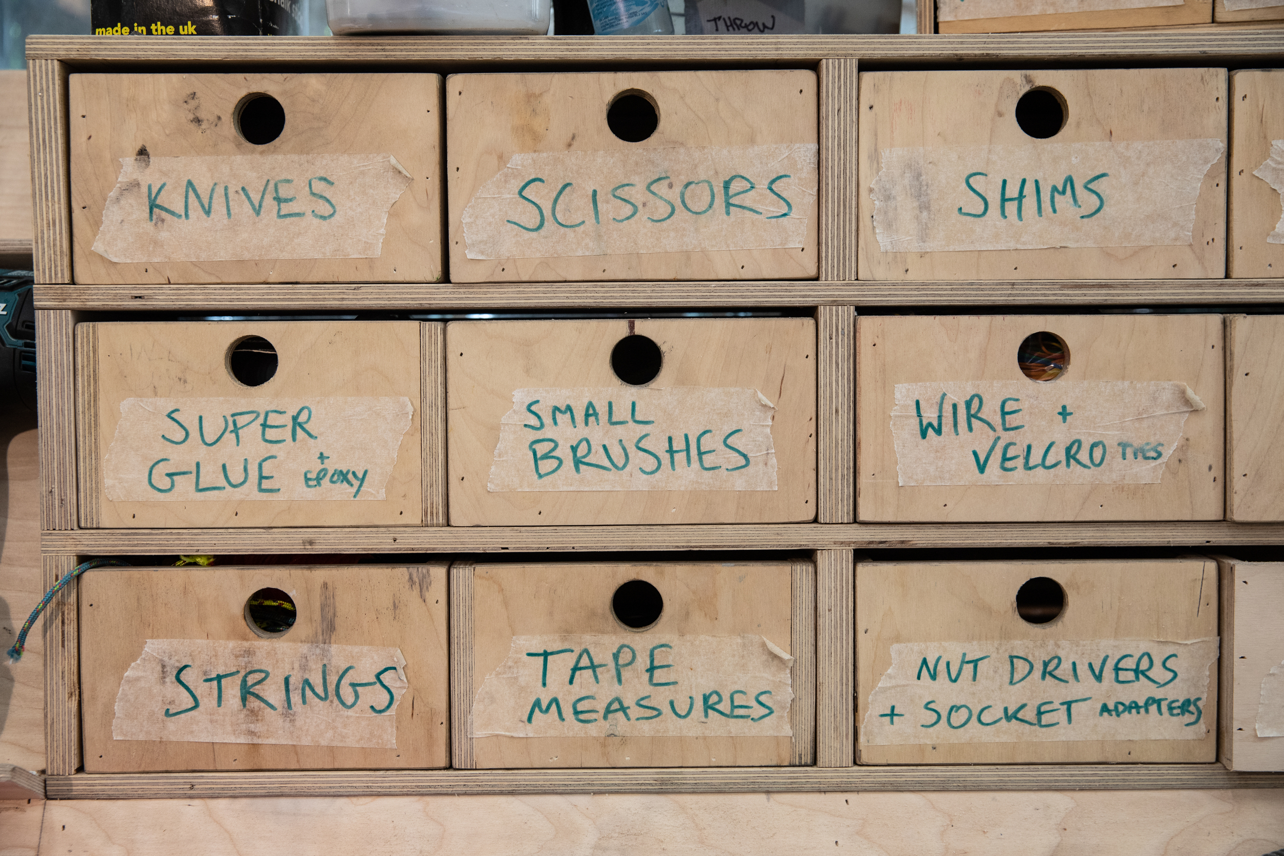 Small wooden drawers with masking tape labels indicating contents including knives, small brushes, tape measures etc