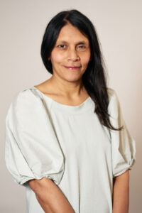 Portrait of Halima Begum, the CEO of Runnymede Trust, with a closed-mouth smile. Her long dark hair contrasts with the cream background and cream top she's wearing.