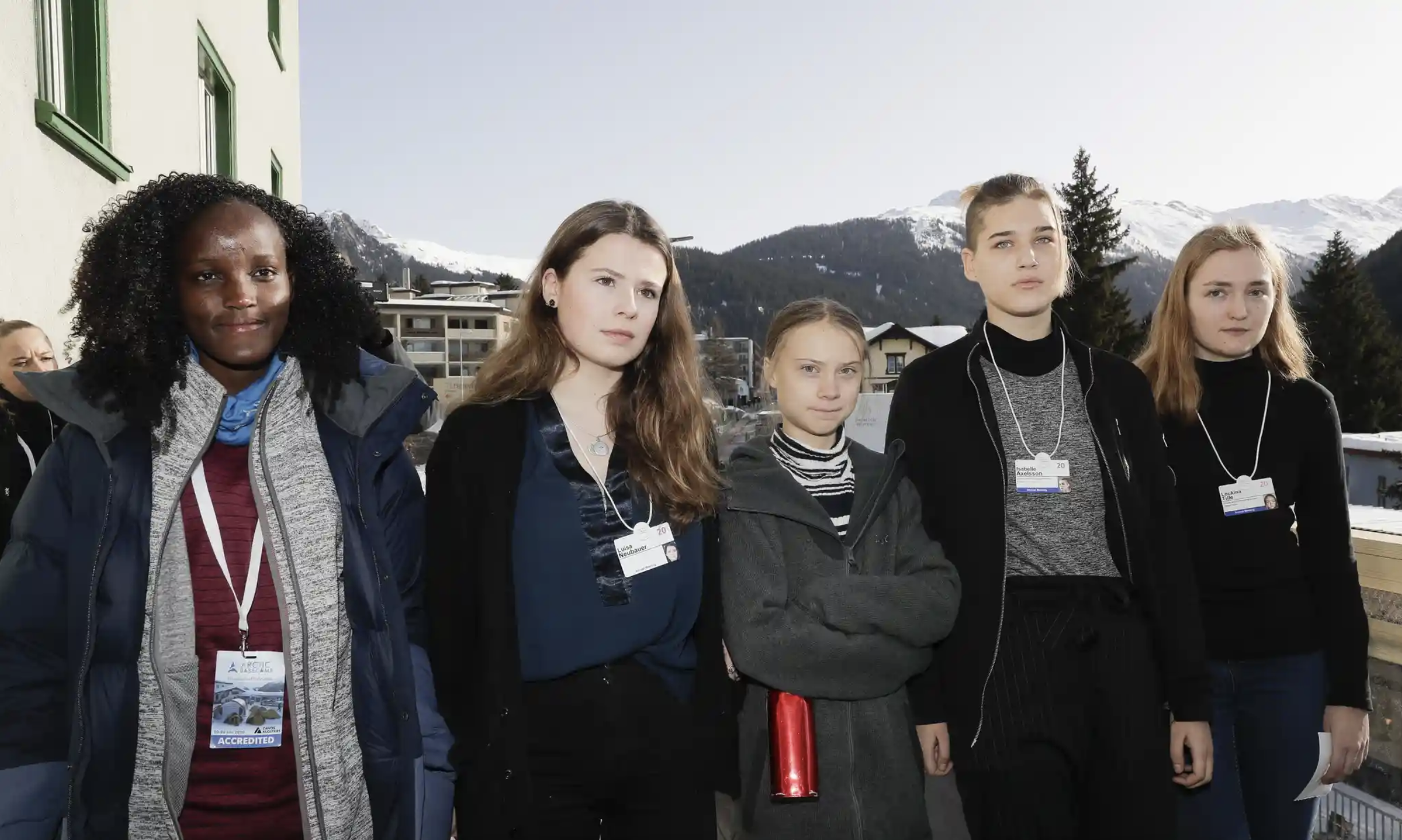 A group of young women stand in a row facing the camera with serious expressions. Four of them are white, and one is Black. They're all wearing smart outdoor clothes and wearing conference lanyards with ID badges.