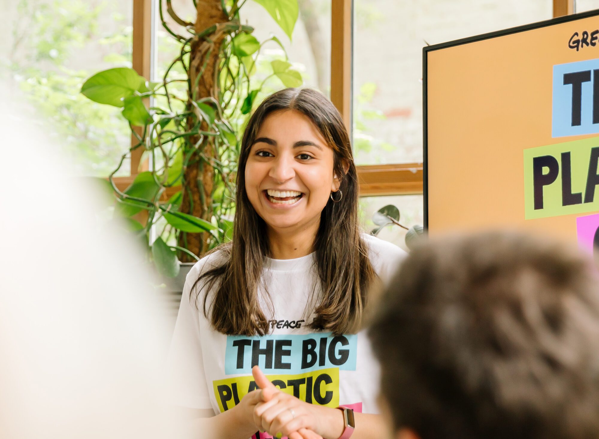 In a bright room with climbing plants in the background, a Greenpeace speaker gives an animated smile as she interacts with her audience during a talk.