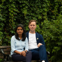 Areeba and Will sitting together on a bench outside, with dense foliage in the background