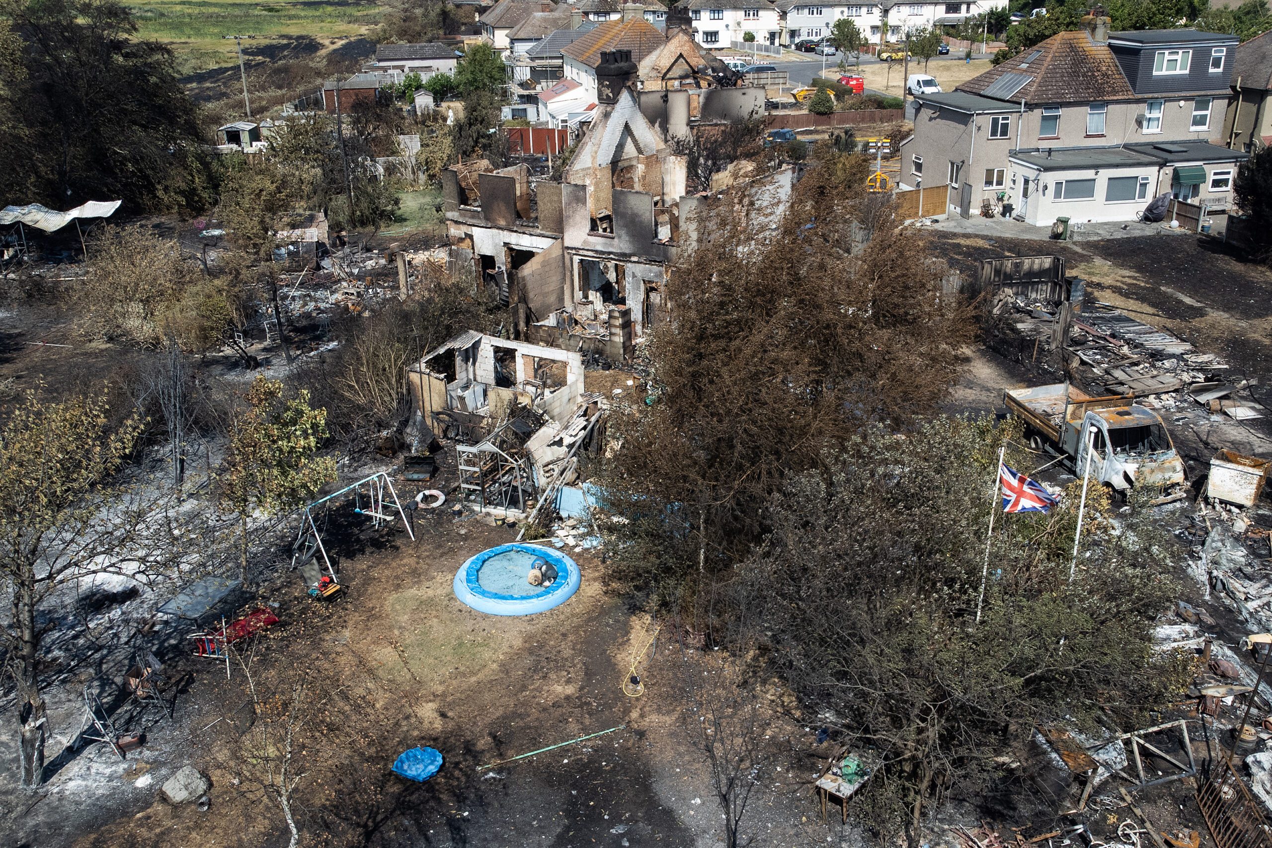 Burnt, skeleton houses destroyed by wildfire. The central house is fallen in and surrounded by dark, ashy ground.
