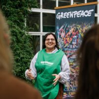A speaker in a green tabard gesticulates as she delivers a talk in front of a Greenpeace banner.