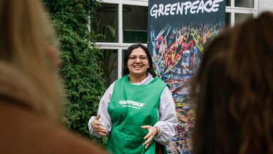 A speaker in a green tabard gesticulates as she delivers a talk in front of a Greenpeace banner.