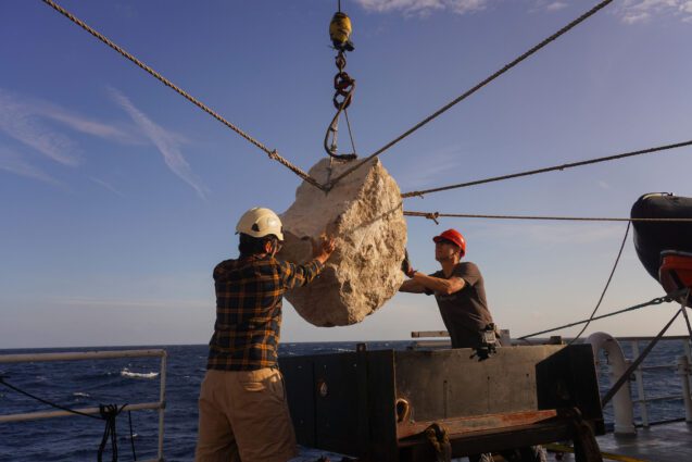 Two people supporting a large boulder being lifted by ropes and pulleys on a ship