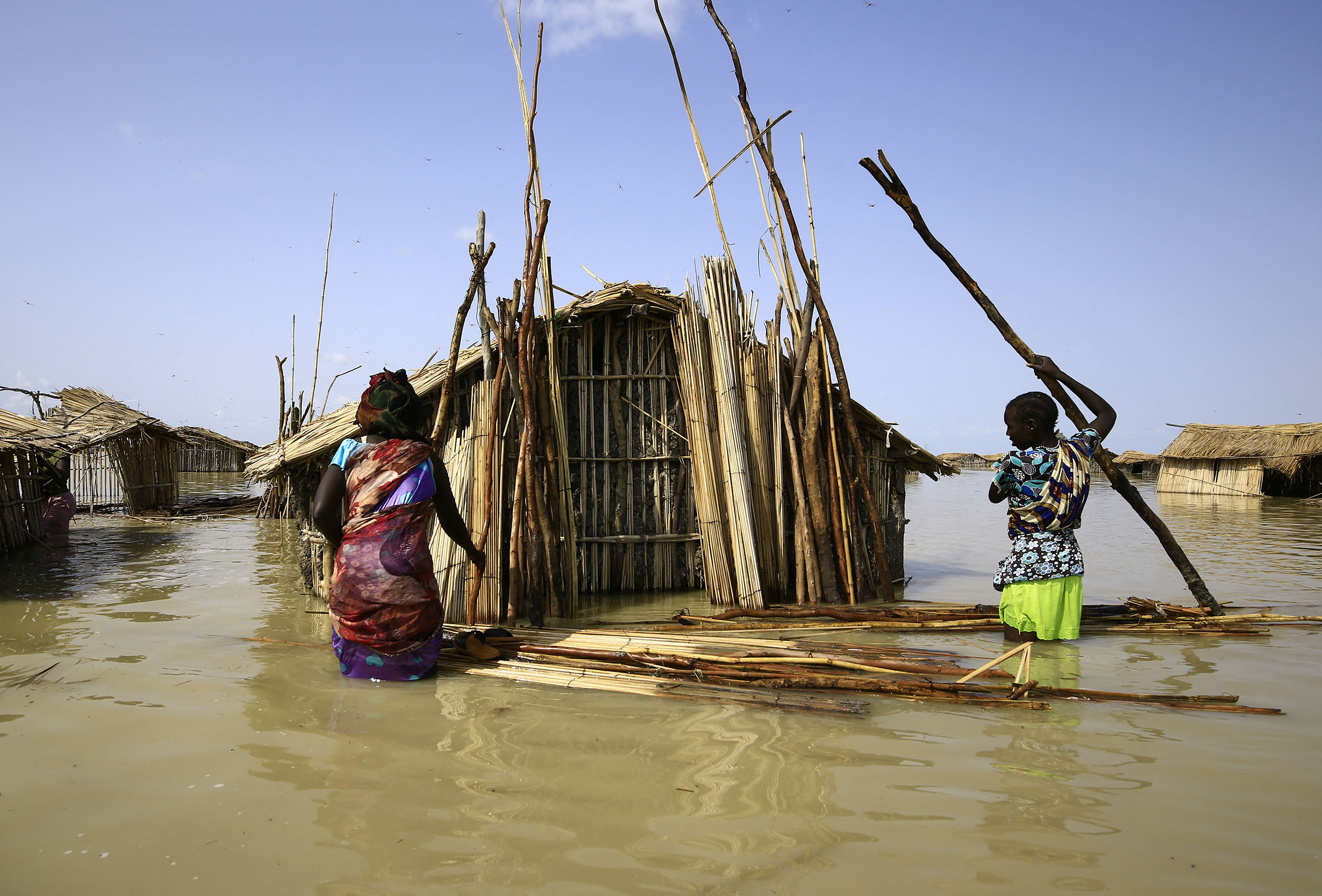 Two South Sudanese women stand in hip-high water next to their damaged home