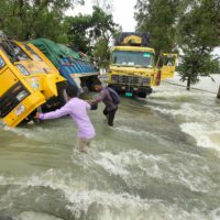 Two men wade through rushing water on a flooded road. They are passing two stranded trucks, one of the has fallen to its side.