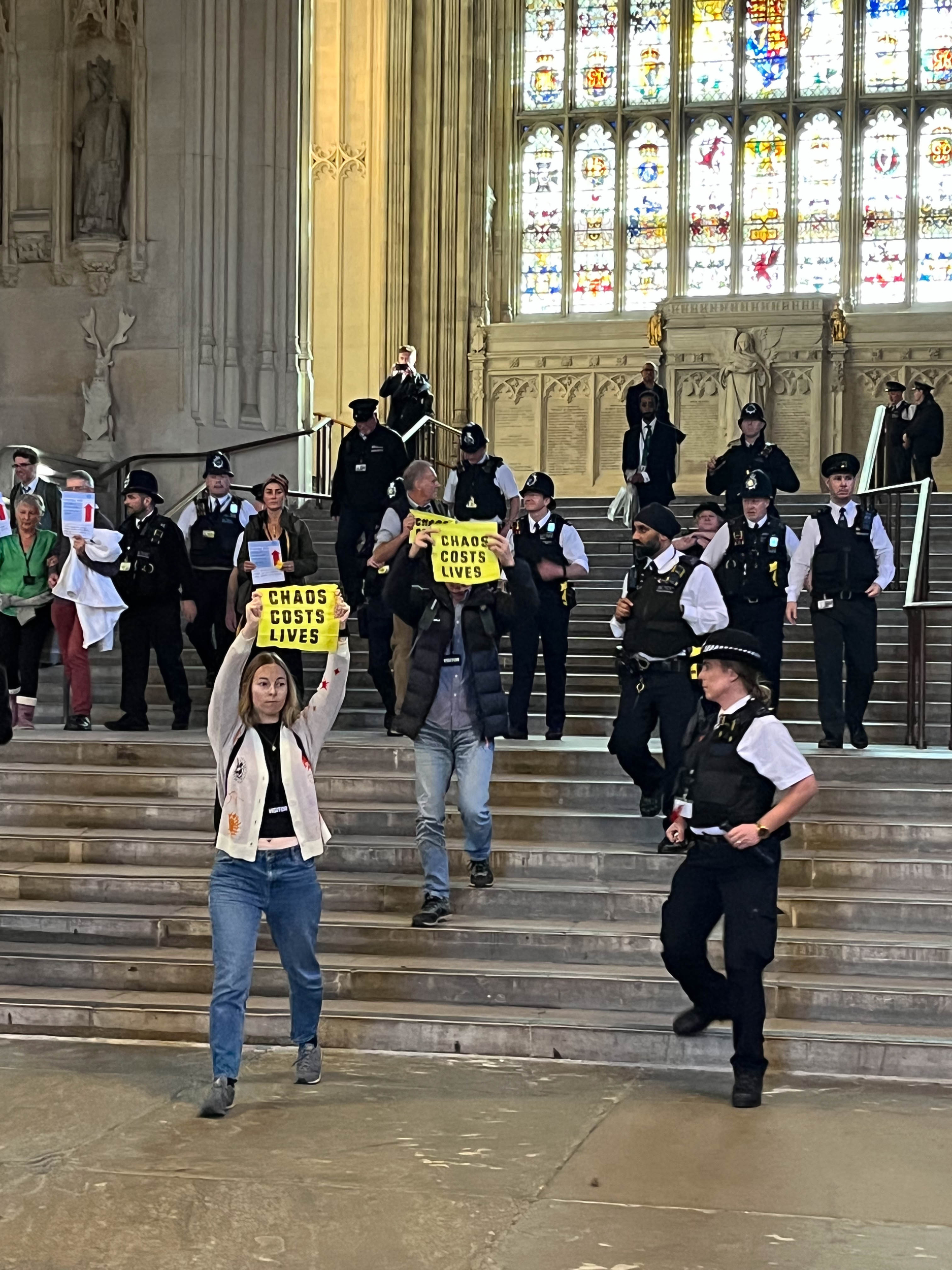 Activists holding 'chaos costs lives' signs while walking down steps by an ornate stained glass window in Parliament