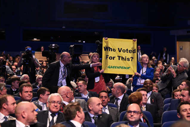 In the audience at the Conservative Party Conference, wo activists hold up a Greenpeace-branded banner reading "Who voted for this?""