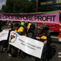 Group of disabled activists protest in a London street. They’re holding banners saying “People before profit” and “Disabled People Against Cuts. Rights not charity”