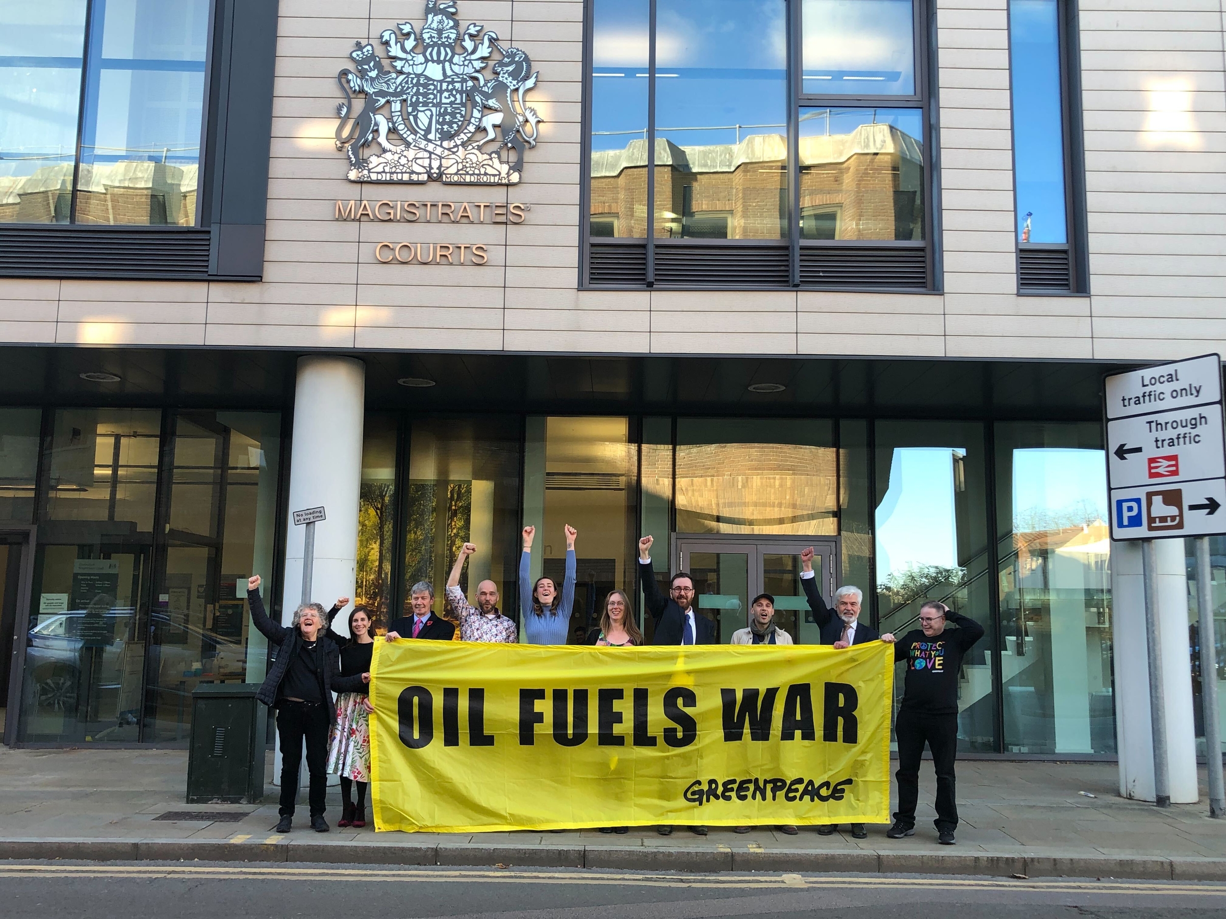 Greenpeace tanker activists celebrate outside court. They thrust their arms in the air and hold a banner reading "Oil fuels war"