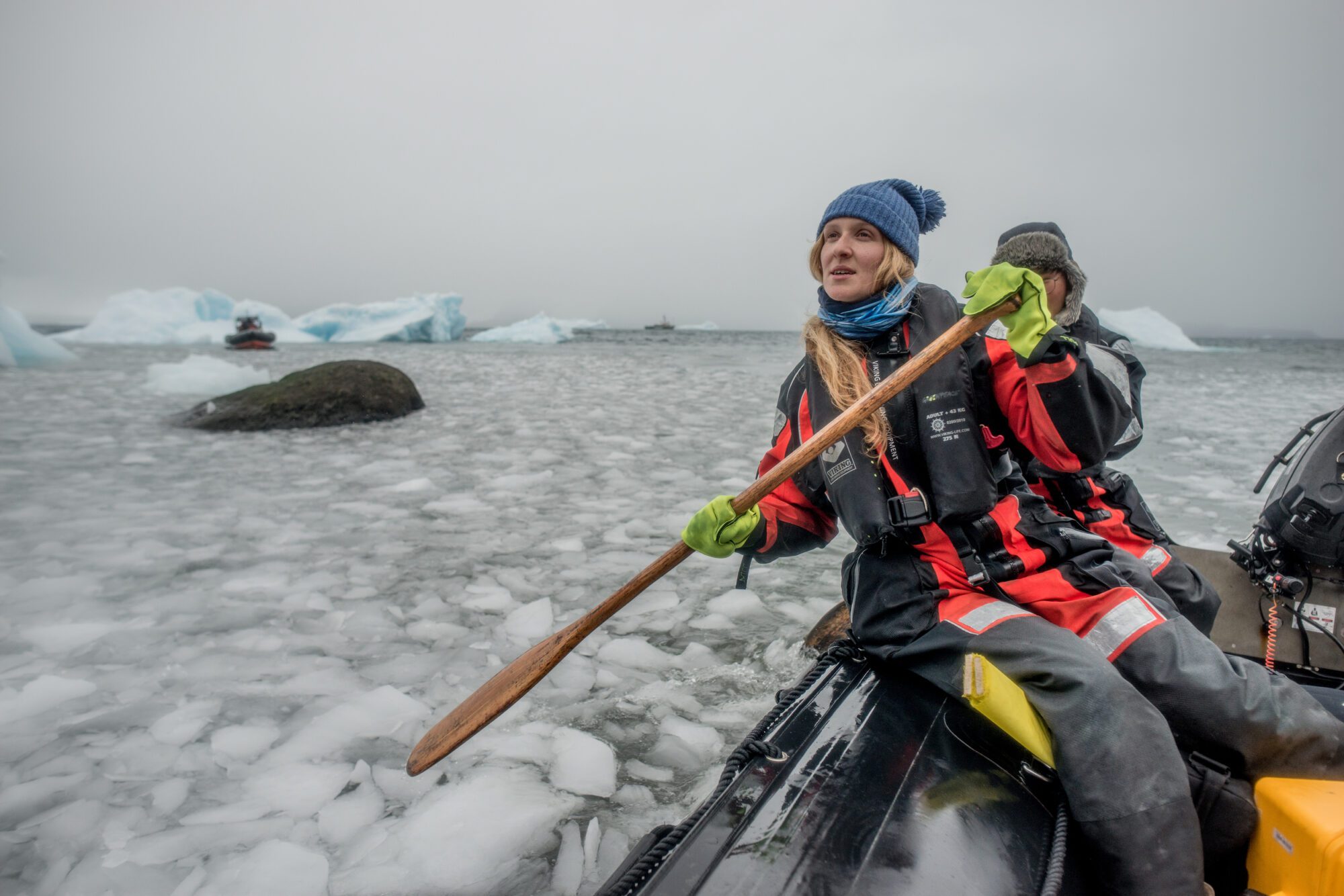 A woman in warm clothing uses a wooden oar to help propel a boat along icy waters.