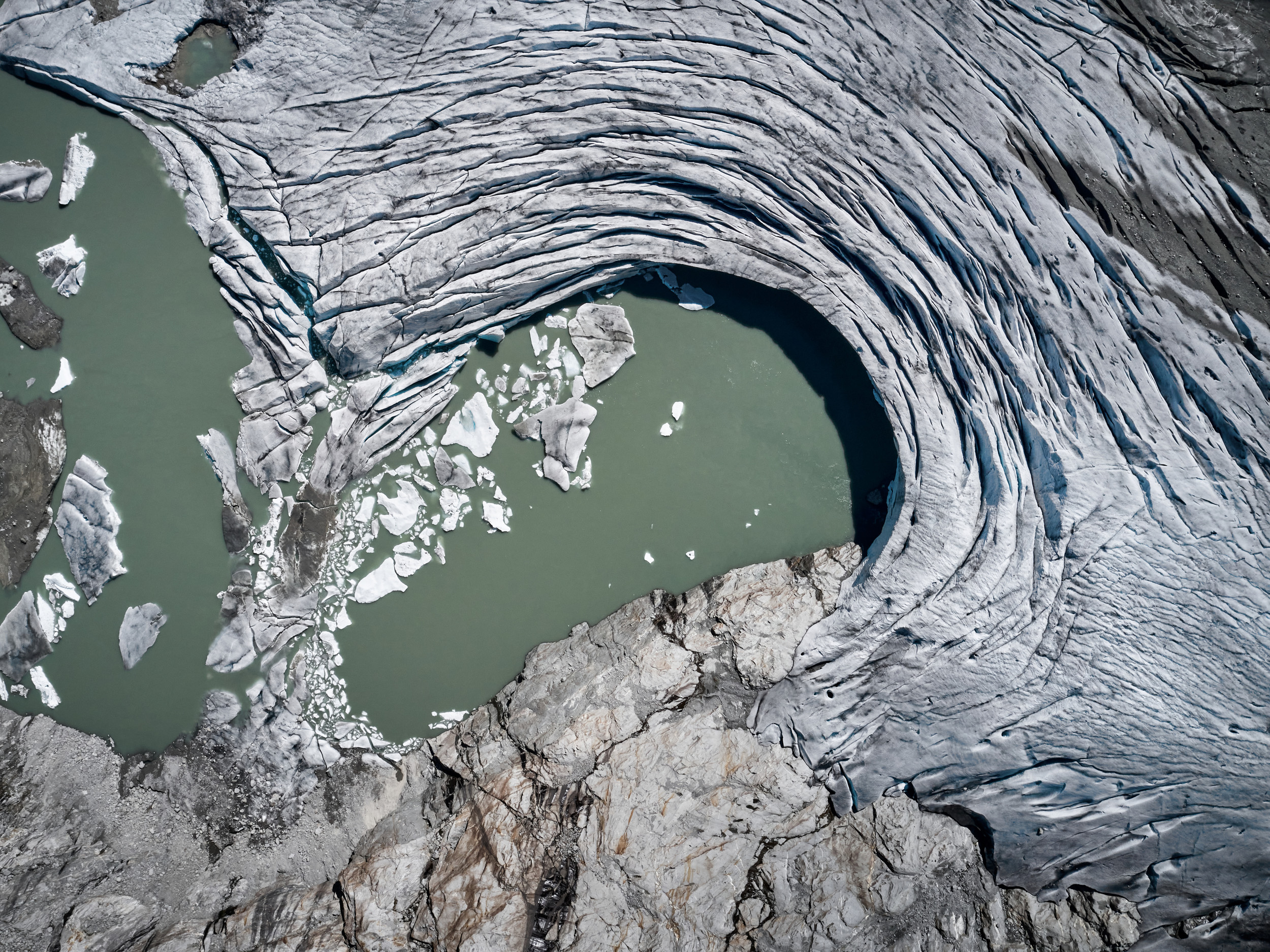 From above, the same wrinkled glacier but the hole has turned into a lake, with sections of glacial ice breaking into the water.