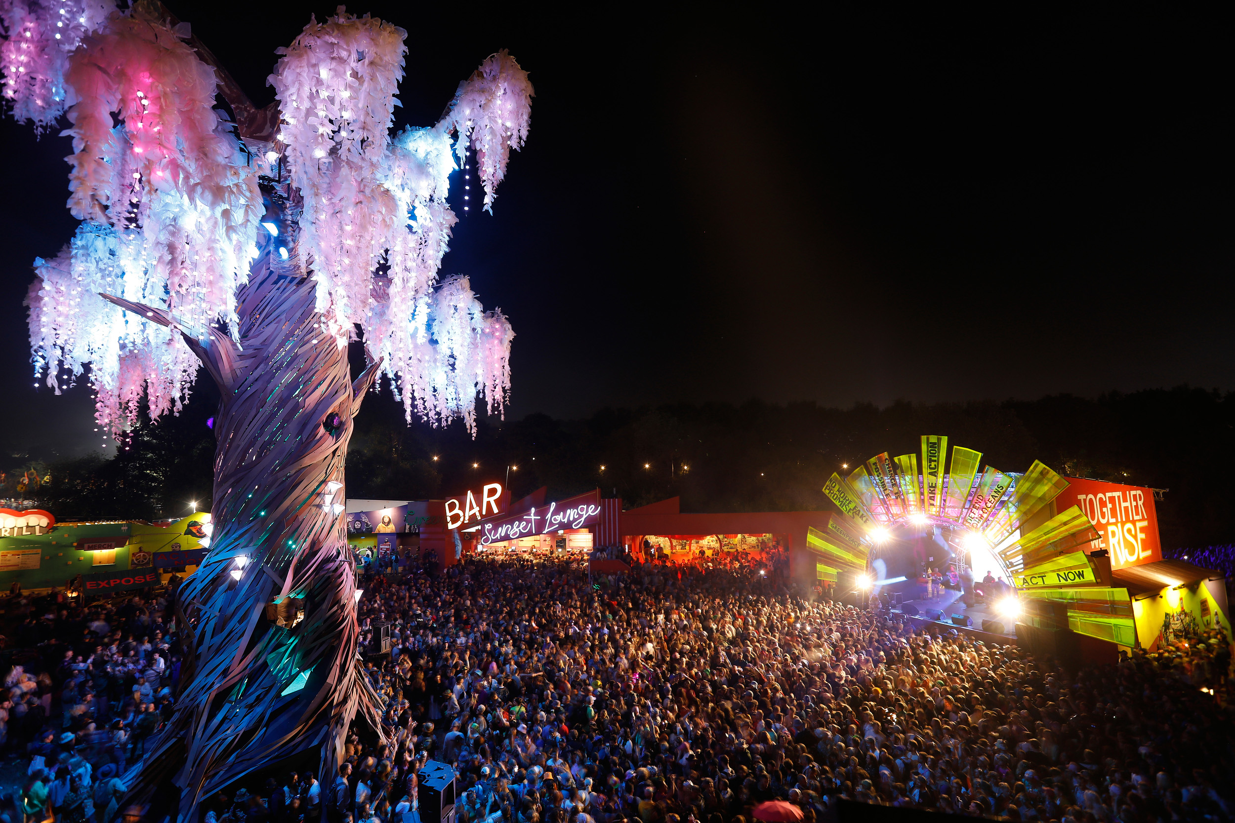 A giant illuminated tree stands proud above a large crowd who are watching live music on a stage at night.