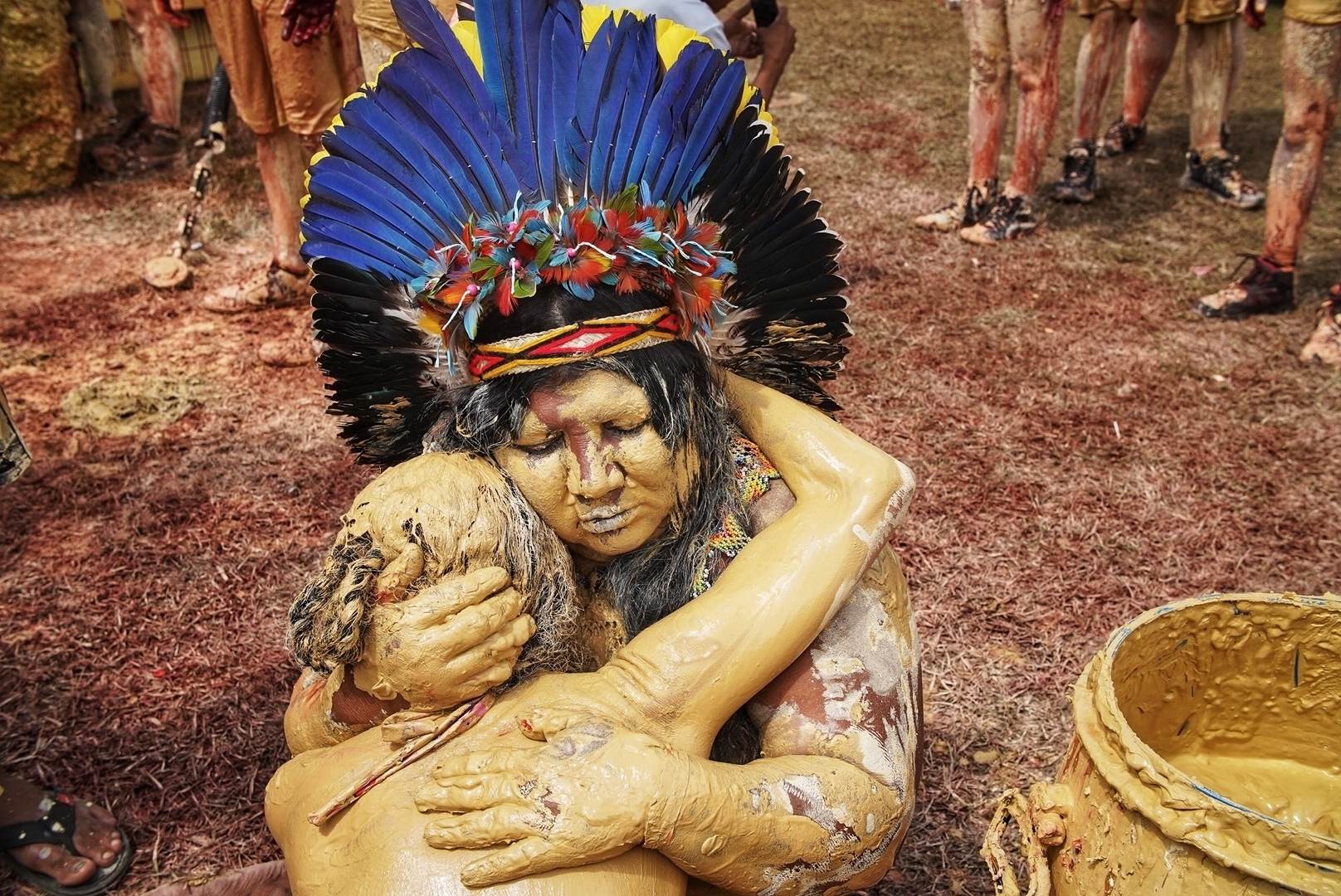 Two people embrace while seated on the ground. Both their bodies are golden and one wears a traditional headdress.