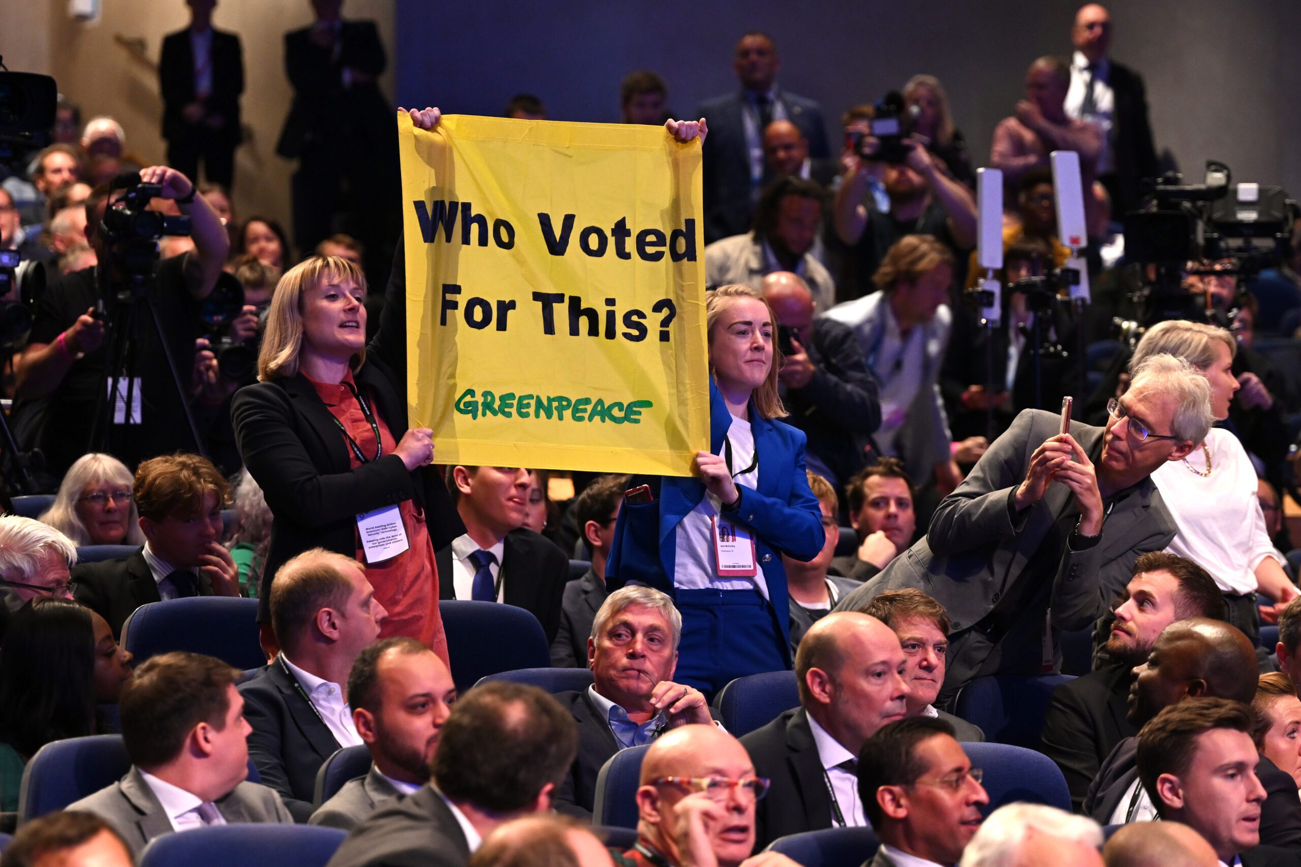Two campaigners standing up in an audience with a banner that reads “Who Voted For This?”