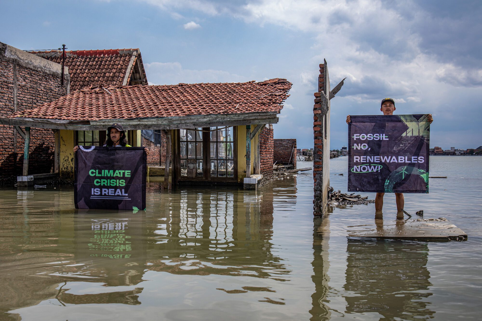Two people stand in floods next to a broken house. Their banners read “Climate crisis is real” and “Fossil no. Renewables now”