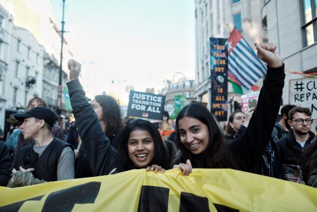 Two protesters raise their fists in solidarity during a march. Behind them, other protesters hold banners saying “Justice transition for all” and “This pale blue dot is all we’ve got”.
