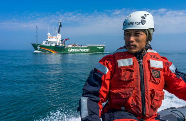 A Greenpeace crew member in a hard hat and red boat suit pilots a small inflatable boat across a still ocean. A Greenpeace ship can be seen in the background.