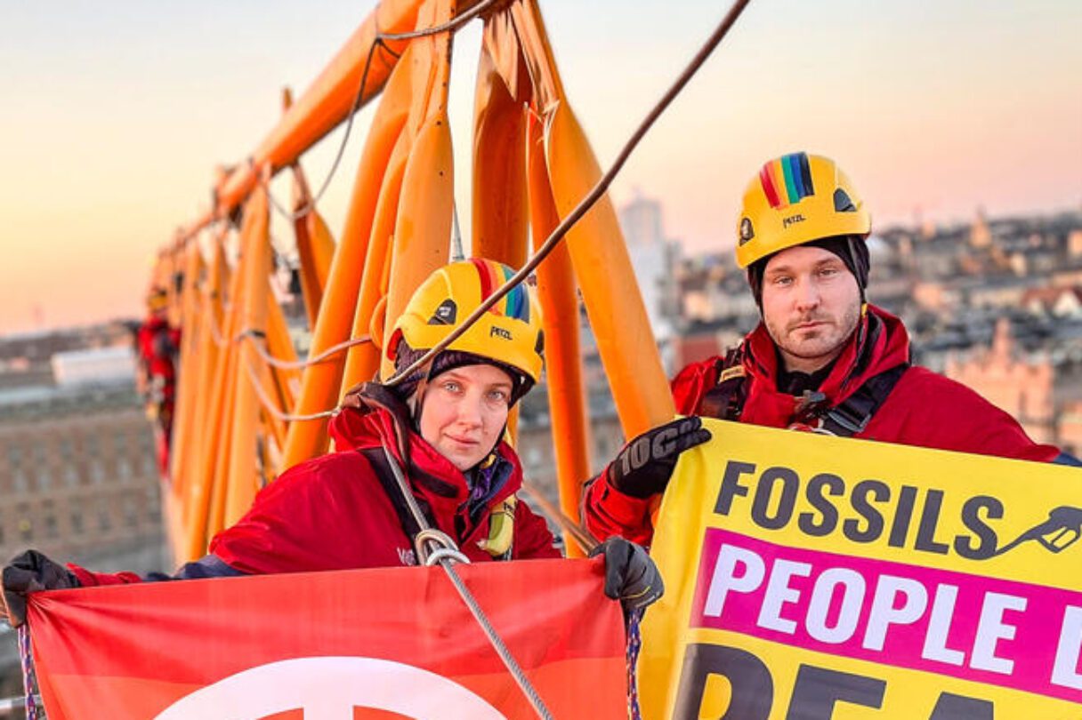 Two climbers wearing helmets and red overalls hold campaign banners on the arm of a crane, high above a beautiful urban sunrise.