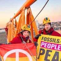 Two climbers wearing helmets and red overalls hold campaign banners on the arm of a crane, high above a beautiful urban sunrise.