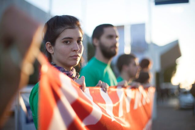 A row of activists hold up a large banner (message not visible). One activist looks into the camera with a determined expression.