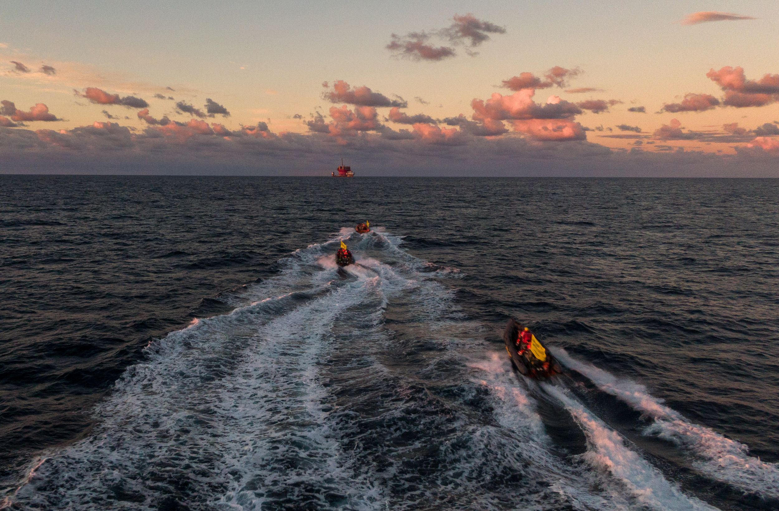 Three boats create white bow waves as they travel toward a distant object at first light.