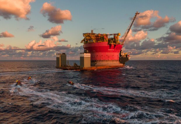 Giant red cylindrical oil platform travels on a ship along the ocean. Activist boats zoom towards it. It looks like David approaching Goliath because they're so small in comparison.