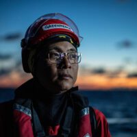 Portrait of Yeb Saño in a Greenpeace hard hat and life jacket against a deep blue and orange sunrise.