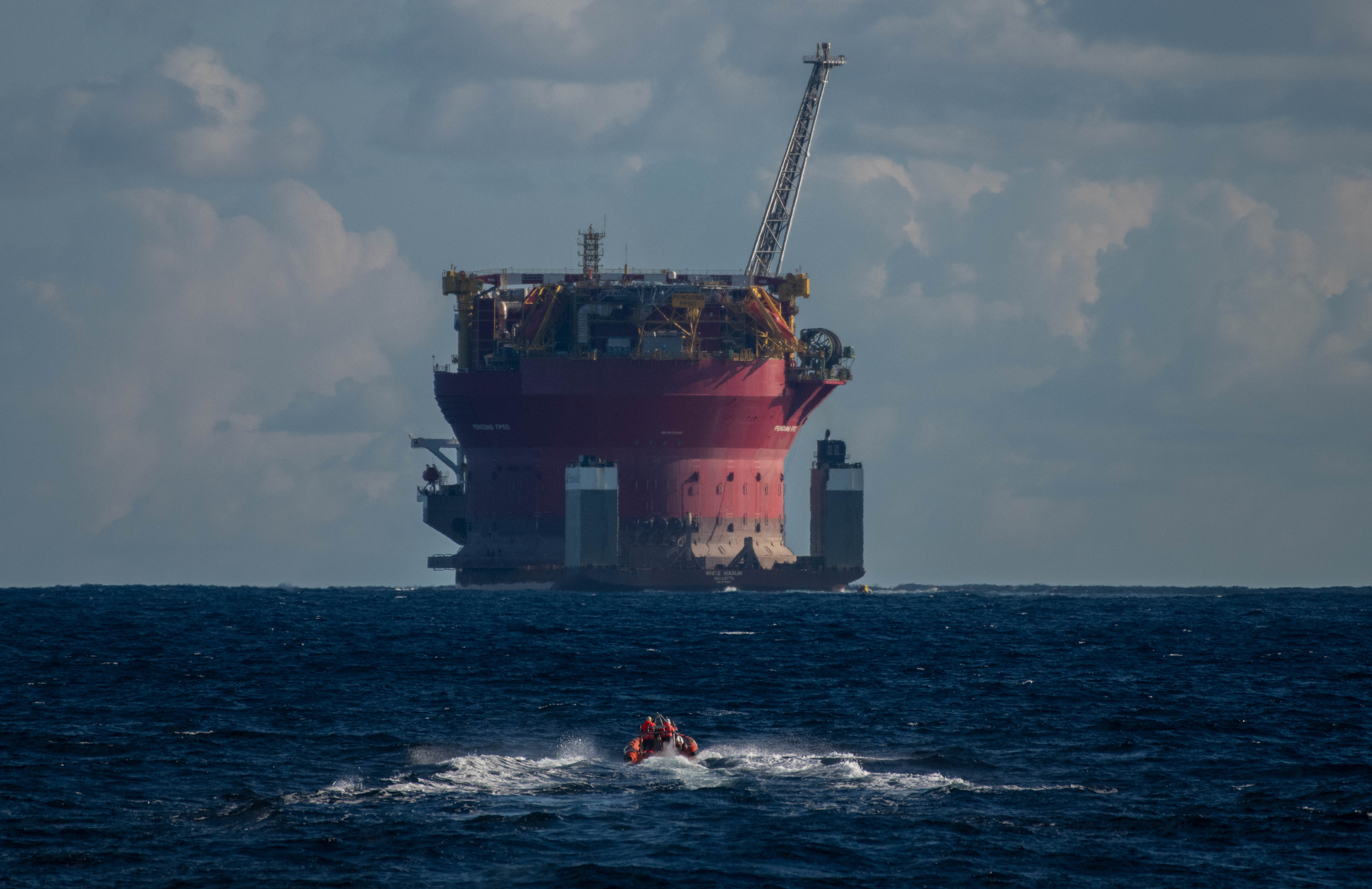 A small boat approaches a giant oil platform in open ocean. The red platform is mounted on a ship, and it looms against the horizon.