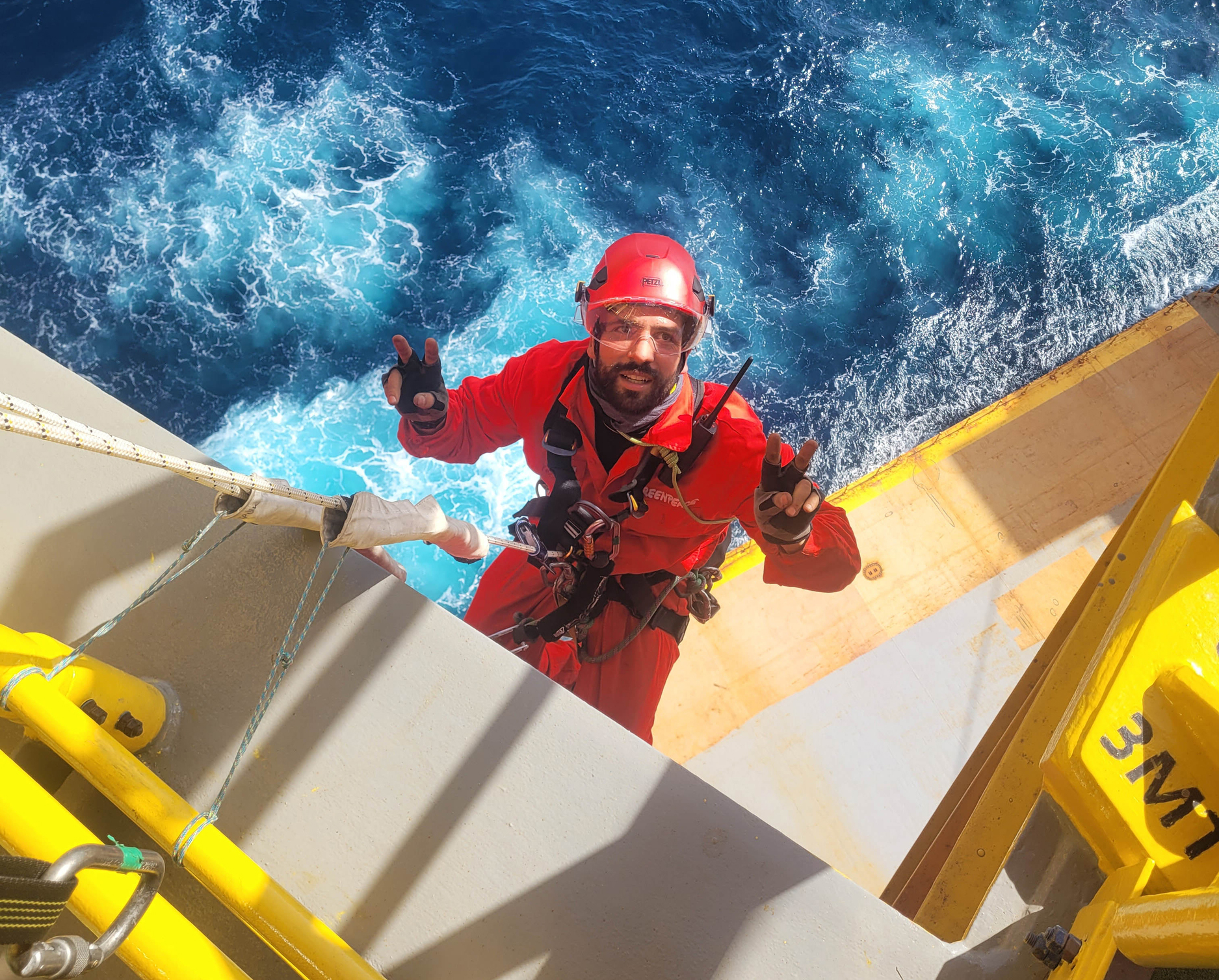 A greenpeace activist in a helmet and red overalls abseiling off an industrial platform over the ocean. He's flashing a peace sign to the camera.