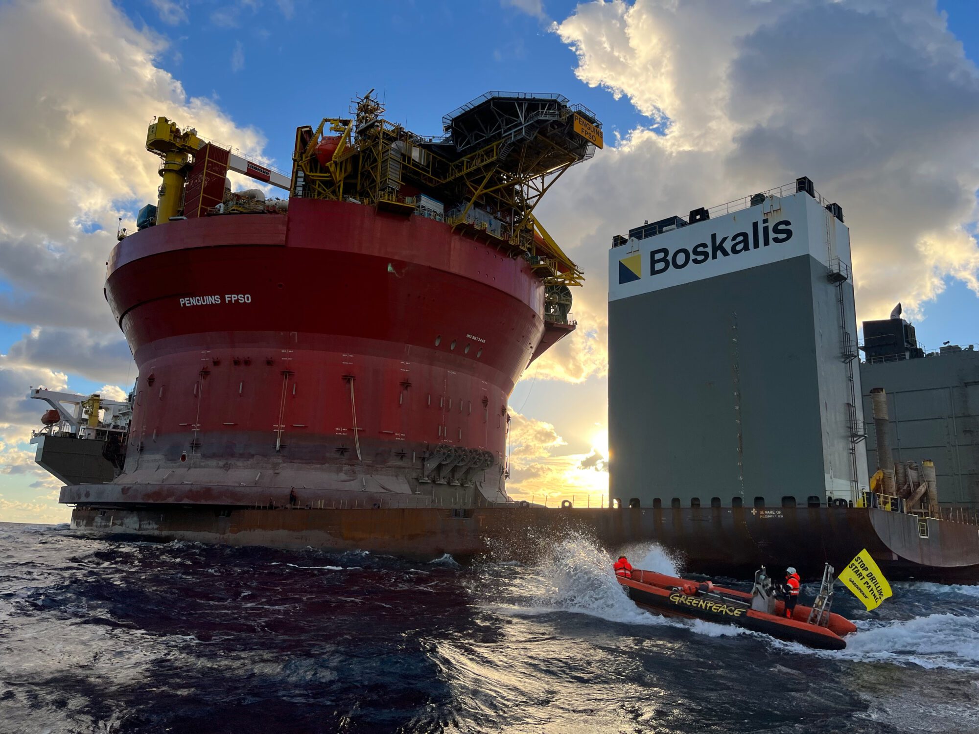 A small inflatable boat with the Greenpeace logo on the side approaches a cargo ship carrying a huge cylindrical oil platform.