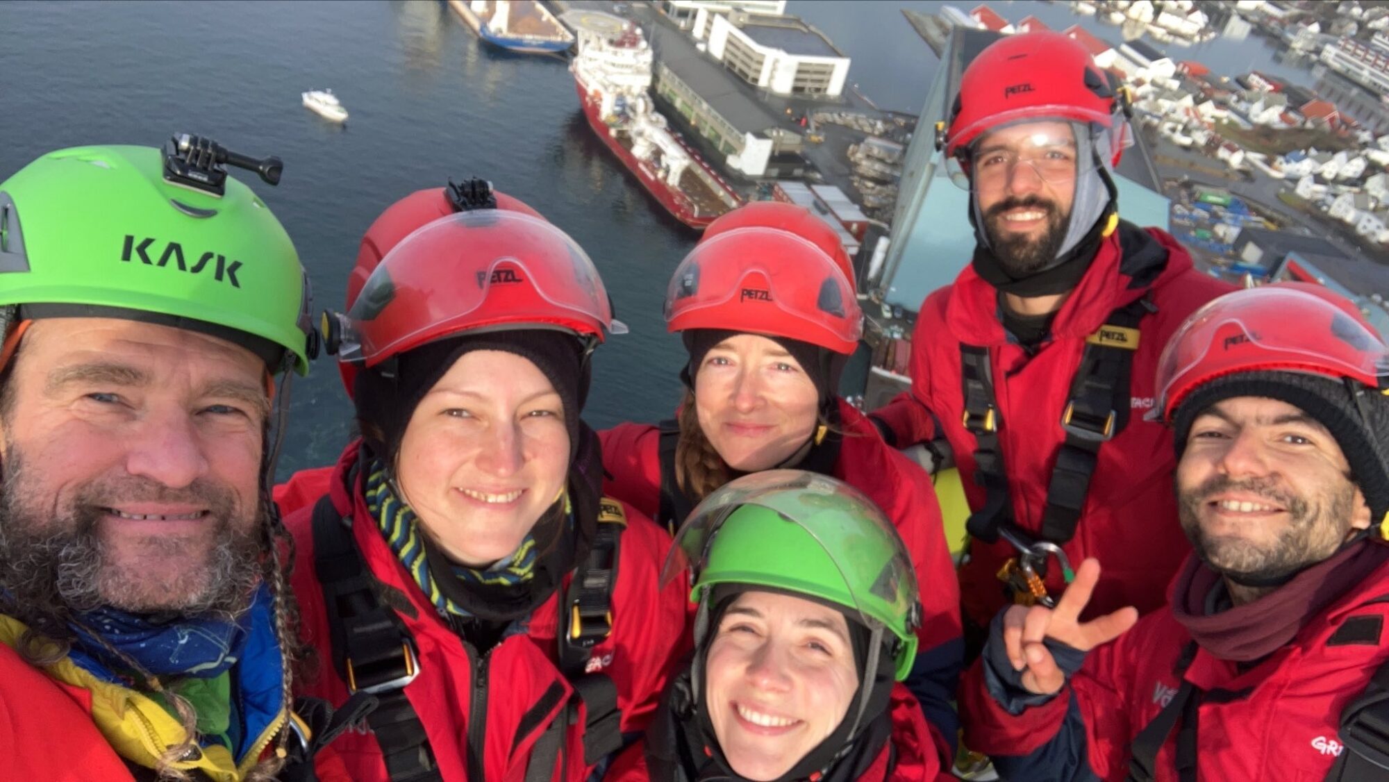 Activists in climbing helmets and red overalls smile for a selfie. Far below in the background, a port city can be seen.