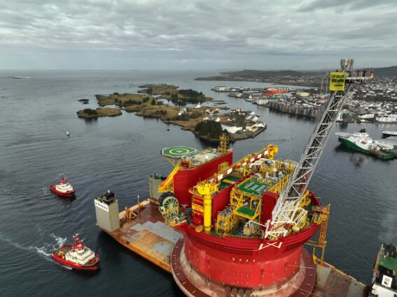 Aerial view of a ship carrying a giant oil platform entering a Norwegian port. The bright red platform dwarfs the surrounding islands and buildings.