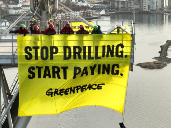 Activists hang a banner reading 'Stop drilling start paying' from the flaring rig of an oil platform, high above a port city.