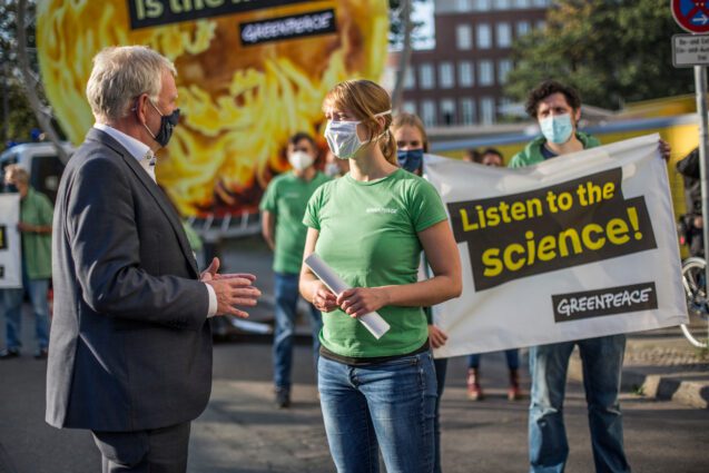 An activist speaks to a person in a suit in front of a banner that says 'listen to the science'