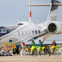 Activists sit around the wheels of a private jet sitting on a runway. More activists on bikes are riding towards them.
