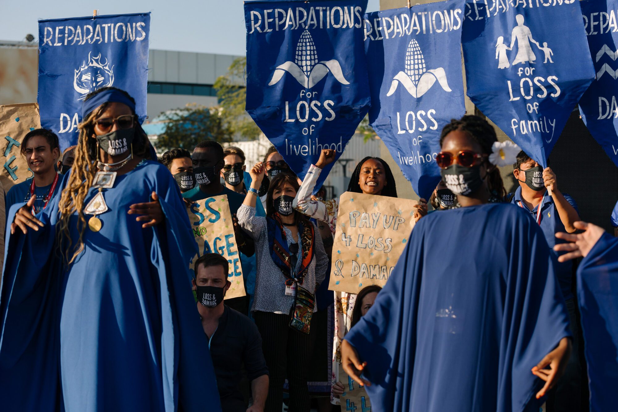 A group of protestors. At the front, woman stand wearing blue gowns and "Loss and damage" emblazoned on face masks. Behind, they hold banners saying "Pay up 4 loss and damage" and "Reparations for loss of livelihood".
