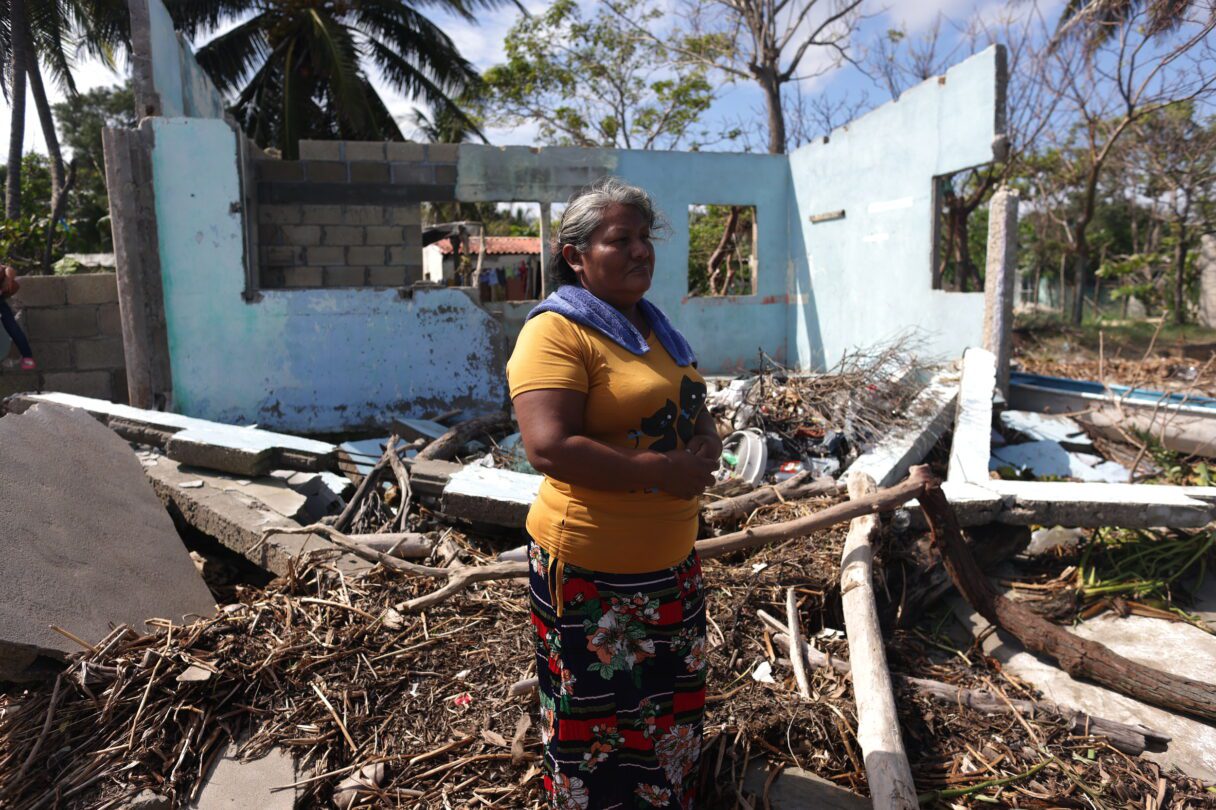 A woman stands in the wreckage of a house. Two upright walls behind her are badly damaged and beneath her feet lie rubble and broken branches.