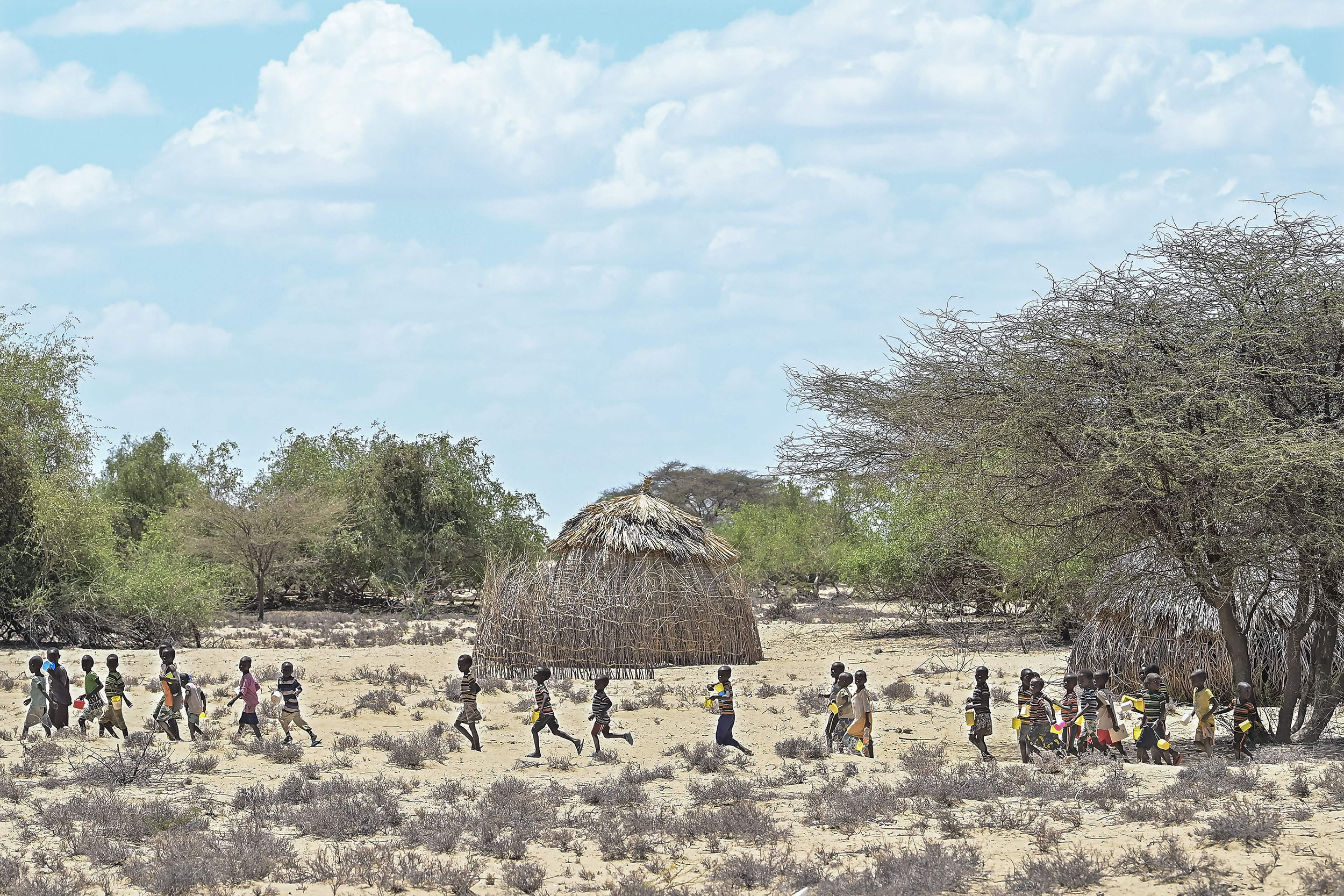 Children run and walk in a line through barren land on a sunny day.