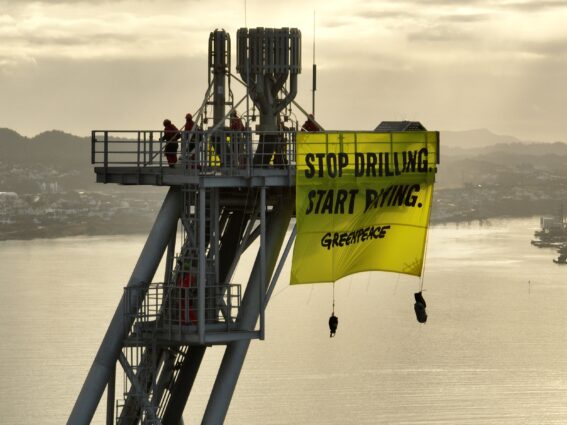 Activists hang a banner reading 'Stop drilling start paying' from the flaring rig of an oil platform, high above a port city.