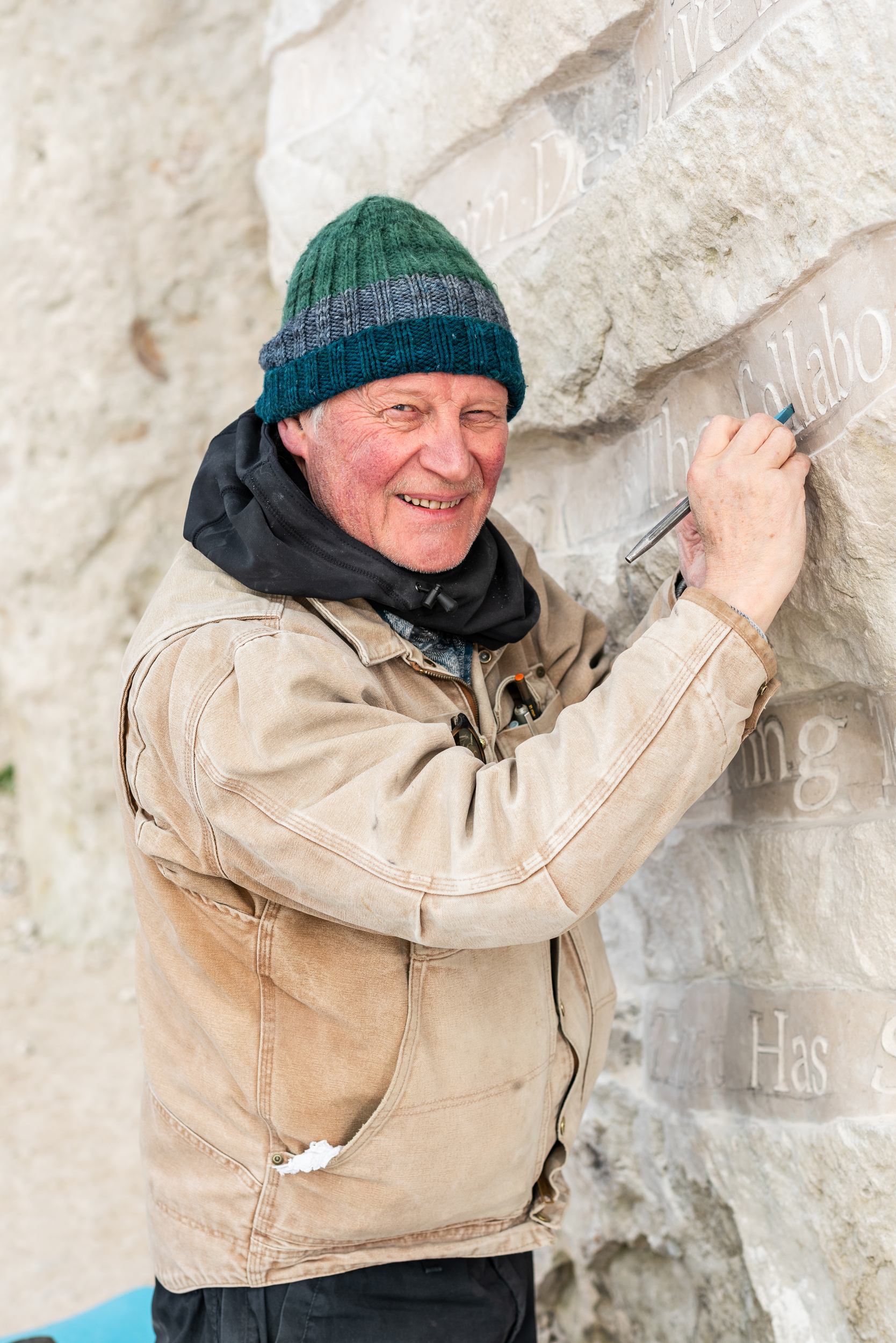 A man smiles and holds tools while standing next to a large stone with carved text on it