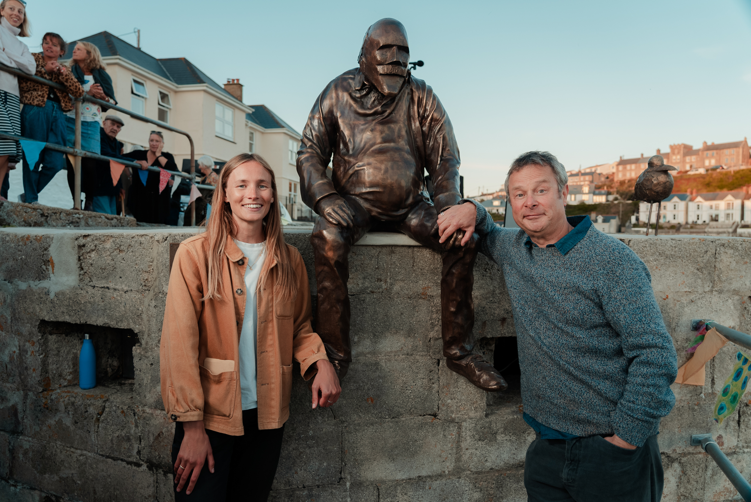 Holly and Hugh pose with a bronze-coloured sculpture of a man sitting with his legs hanging over the edge of a concrete surface. The sculpture hunches slightly forwards and squints into the distance.