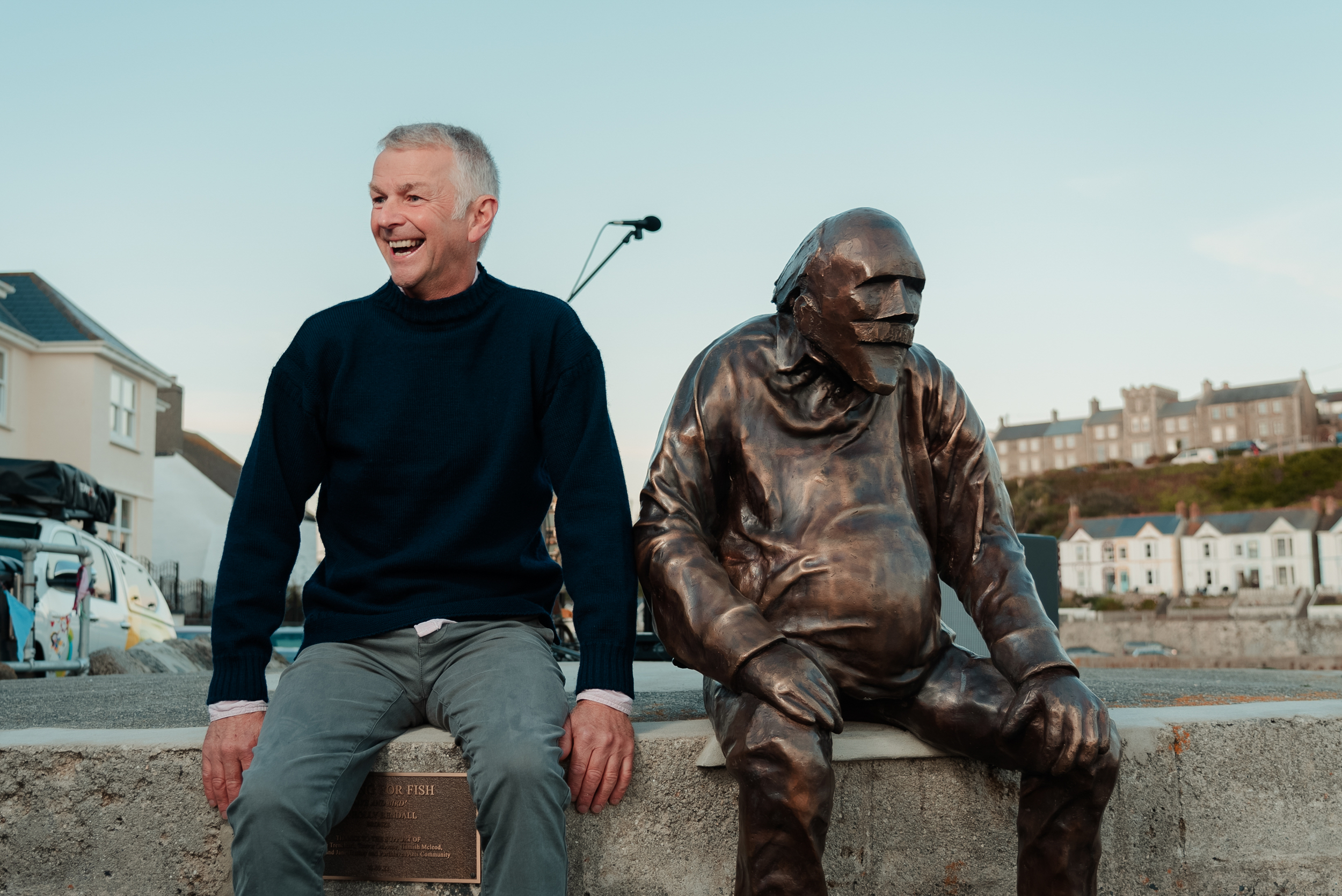 Jeremy smiles while he sits next to a bronze sculpture of a sitting man looking out into the distance.