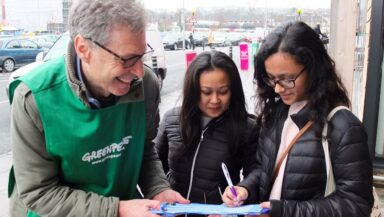 A smiling Greenpeace volunteer helps two members of the public fill in a signup form on a clipboard.