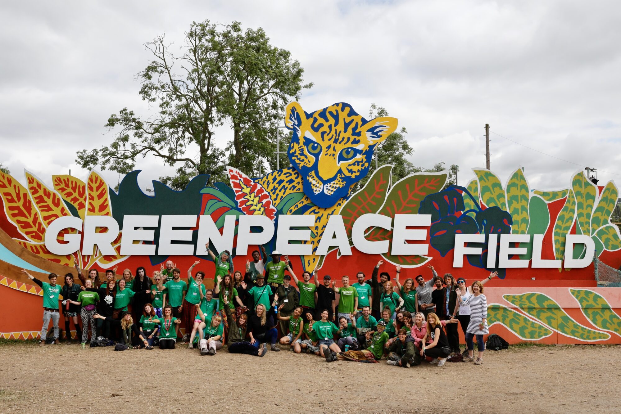 A group of about 30 people in Greenpeace t-shirts stand in front of a giant display board that says Greenpeace Field with colourful jungle designs