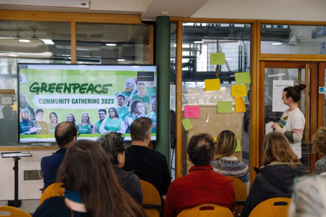 A crowd of people from beind, they are sat looking at a speaker in front of a galls wall with post-its and a screen reading "Greenpeace Community gathering 2023"