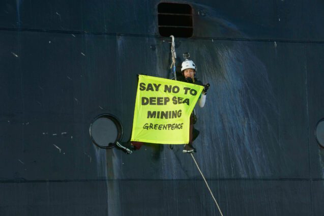 Activist hanging from the side of a ship in climbing gear holding a banner saying say no to deep sea mining