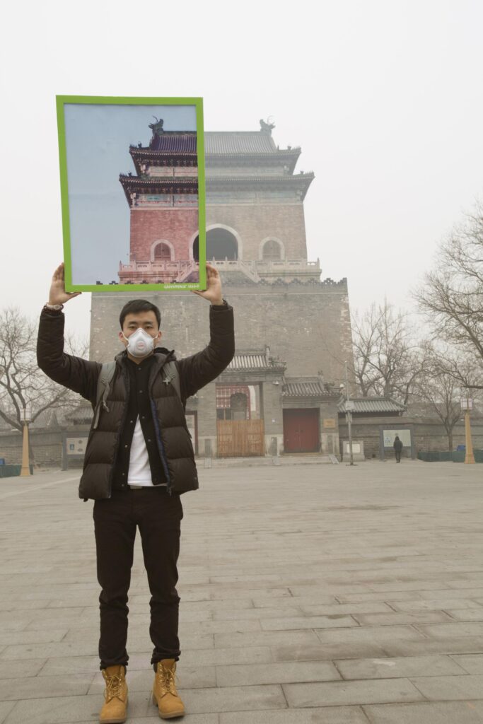 Greenpeace activist in a face mask holds up a large placard with a photo of the corner of the Chinese-roofed building he's standing in front of showing the building with blue sky in its background. The area he is standing in is hazy and the sky is white; the building in the placard photo is red but behind him the building is obscured by haze and is brown/grey. The placard image of the corner is perfectly aligned with the building behind with the air quality/colours the only difference.